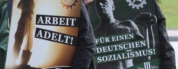 Auf dem Bild sind zwei Aktivisten der Partei Der Dritte Weg zu sehen, die Kundgebungsplakate mit den Aufschriften "Arbeit adelt" und "Für einen deutschen Sozialismus!" halten. Auf dem Bild sind zwei Aktivisten der Partei Der Dritte Weg zu sehen, die Kundgebungsplakate mit den Aufschriften "Arbeit adelt" und "Für einen deutschen Sozialismus!" halten.